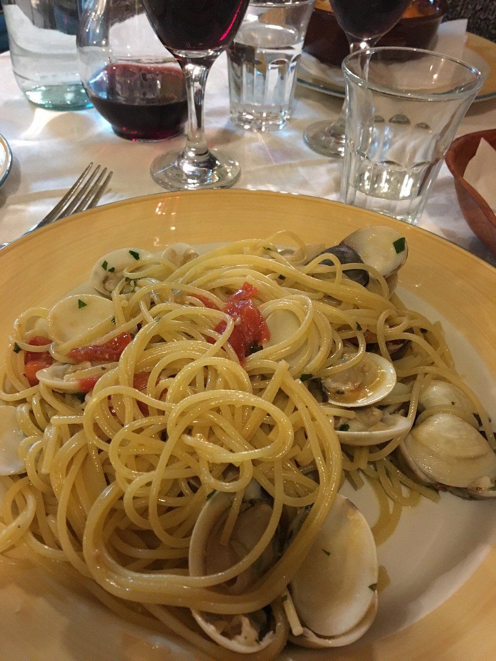 Un piatto di spaghetti con vongole e pomodorini su un tavolo di ristorante, con bicchieri di vino e acqua sullo sfondo.