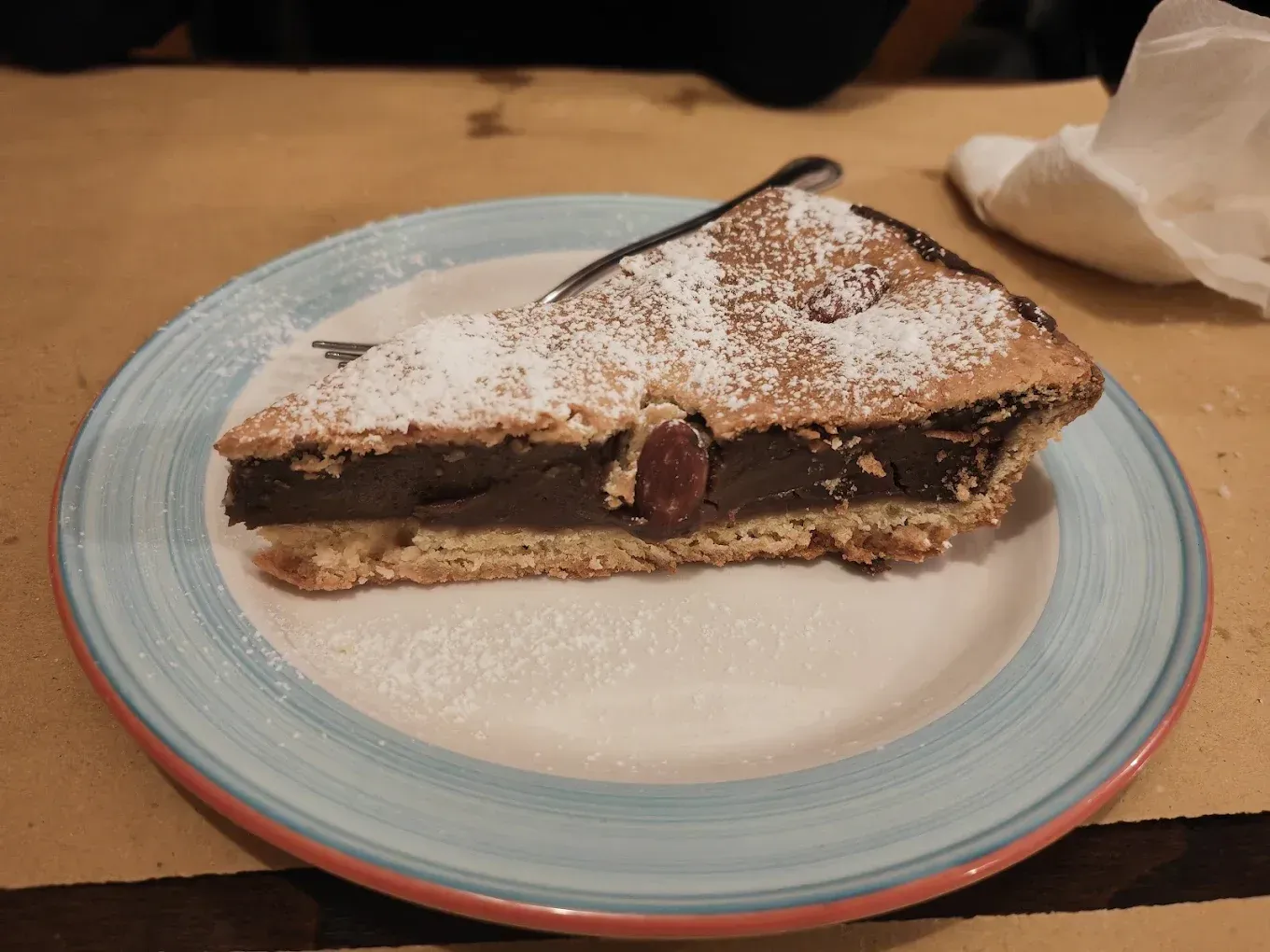 Una fetta di torta al cioccolato spolverata di zucchero a velo su un piatto con il bordo blu a un tavolo