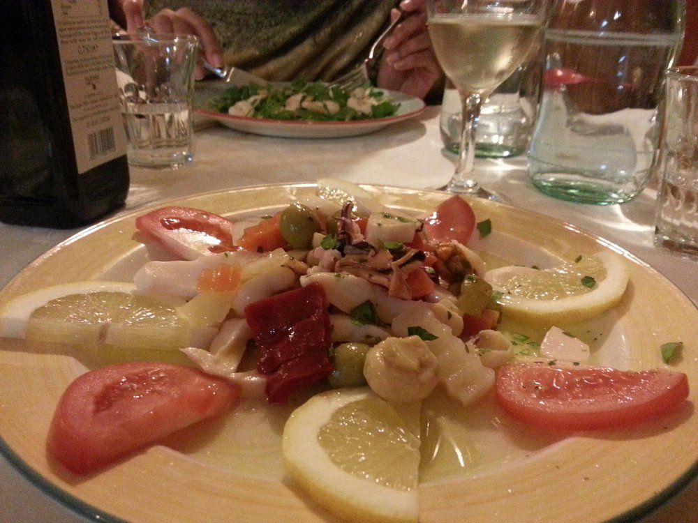 Antipasto di frutti di mare con spicchi di limone su un tavolo di ristorante, con bicchieri di vino e commensali sullo sfondo.