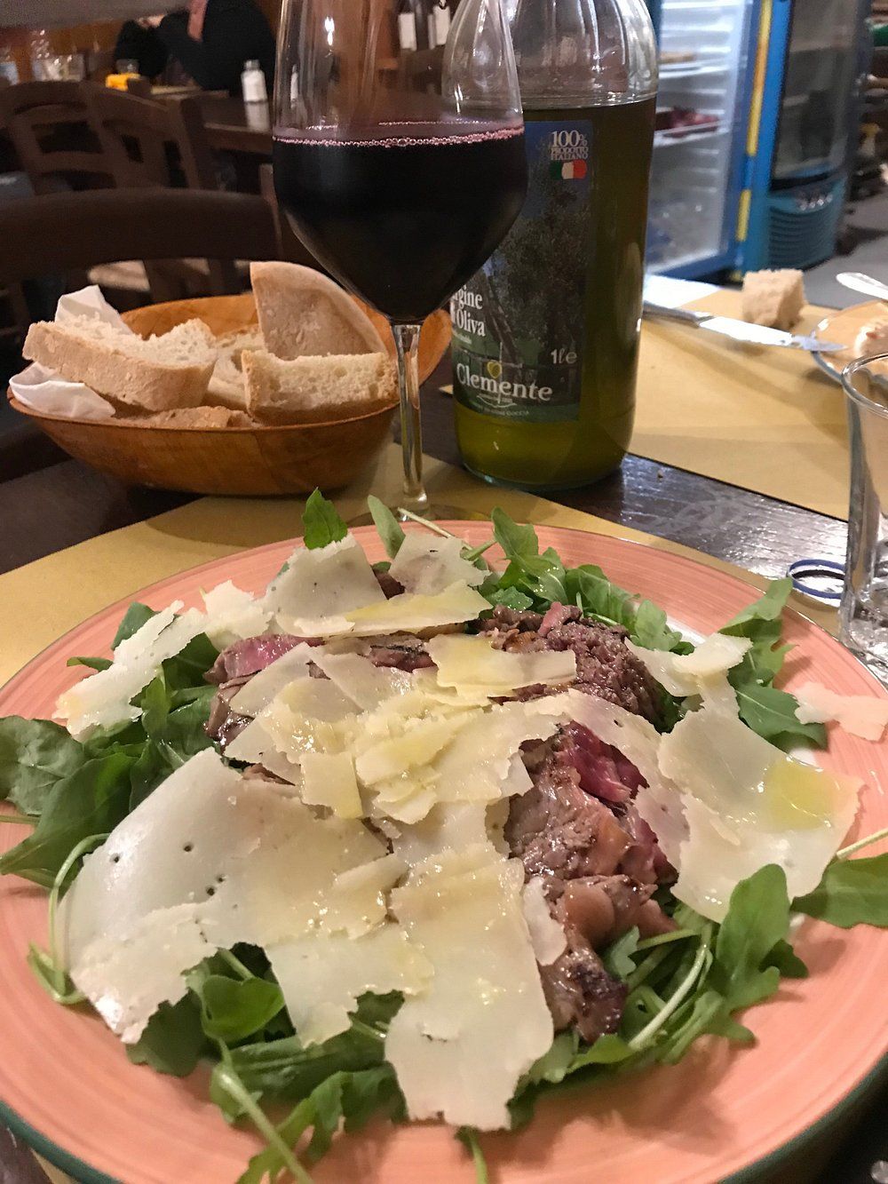 Insalata con scaglie di formaggio su un tavolo di ristorante, con pane e un bicchiere di vino sullo sfondo.