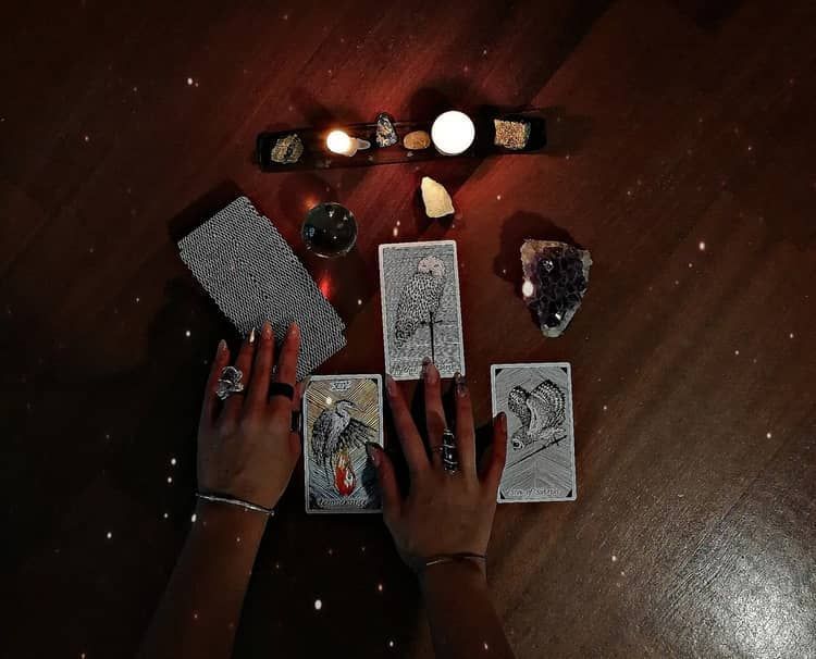 Hands touching Tarot cards on a wooden table with crystals, candles, and a deck. Dark, mystical setting.
