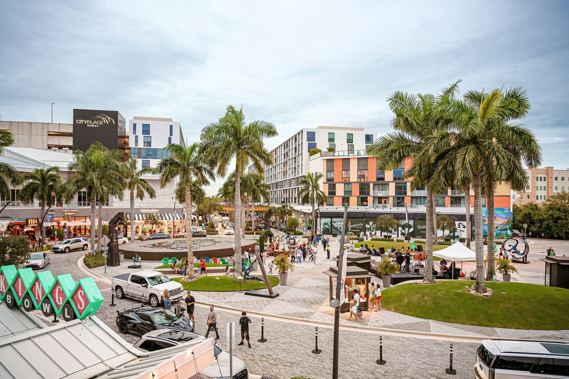Plaza with buildings, palm trees, and people. Gray brick paving, vehicles, and a cloudy sky.