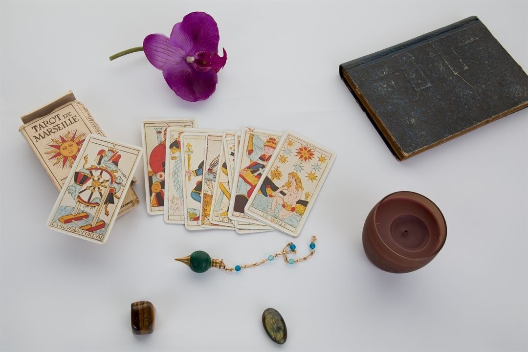 Tarot cards, purple orchid, small book, beads, and candle on white surface.