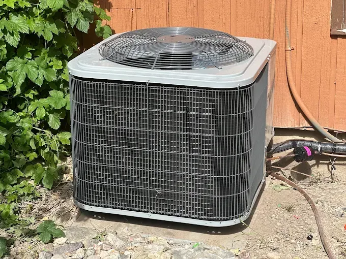 An air conditioner is sitting on the ground next to a bush.