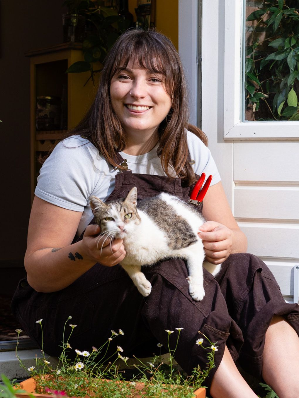 Een vrouw in een bruine overall glimlacht terwijl ze buiten een wit-grijze kat op haar schoot houdt.