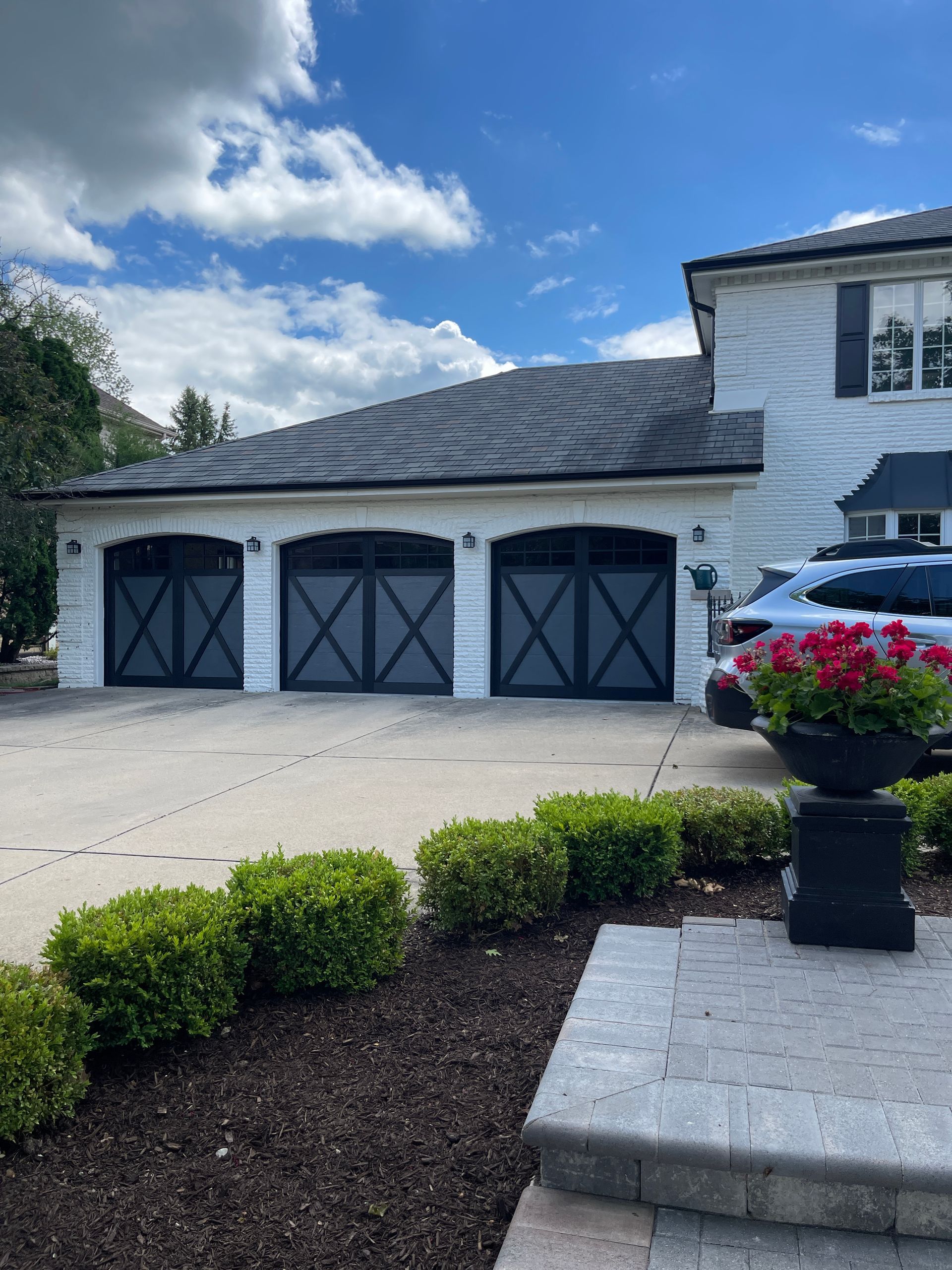 Three-car garage with black doors and white trim. Exterior view with bushes, a walkway, and a car. Blue sky.