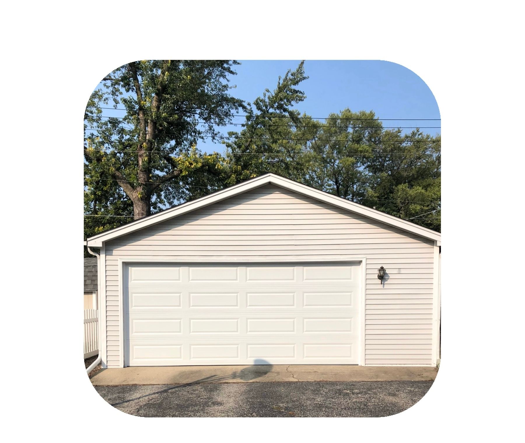 Garage with white door, light siding, and trees in the background under a blue sky.