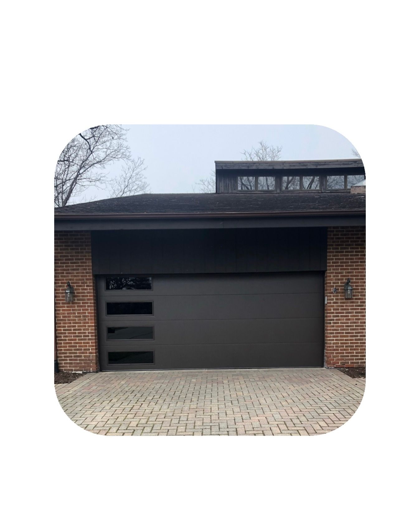 Dark gray modern garage door with windows, flanked by brick, over a paver driveway.