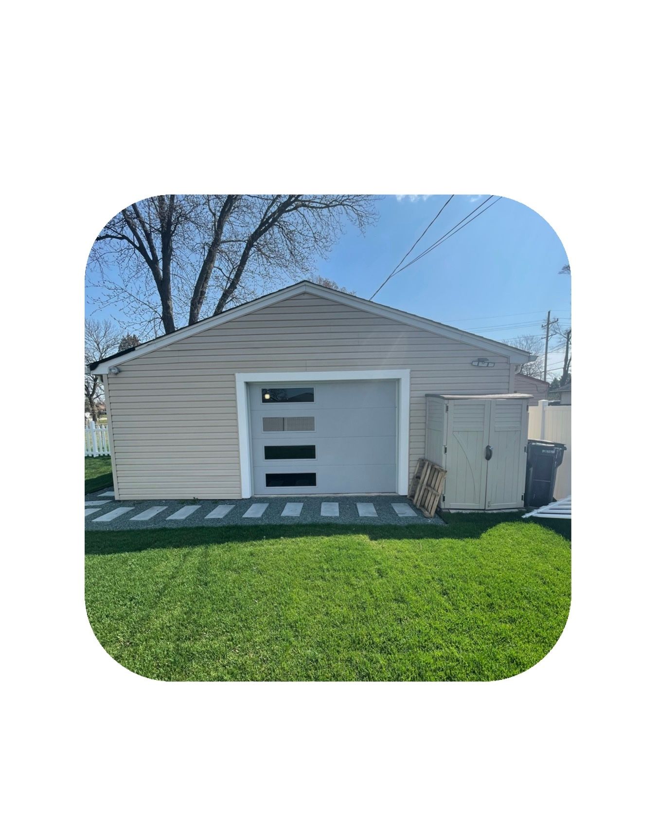 Tan detached garage with gray door and accent windows, surrounded by green grass and a blue sky.
