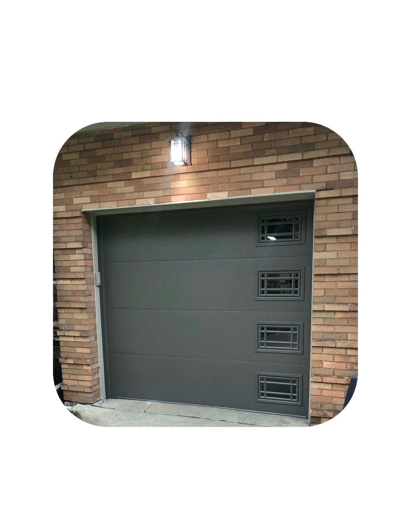 Gray garage door with four windows, in front of a brick building, lit by an overhead light.