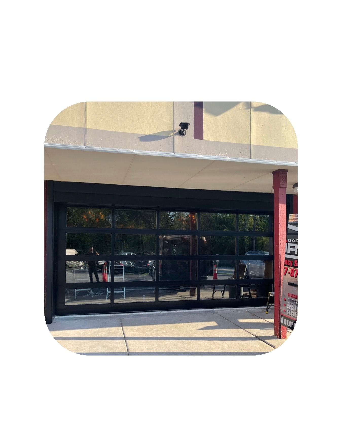 Storefront with black-framed, glass garage door open. Beige building with red columns and a security camera.