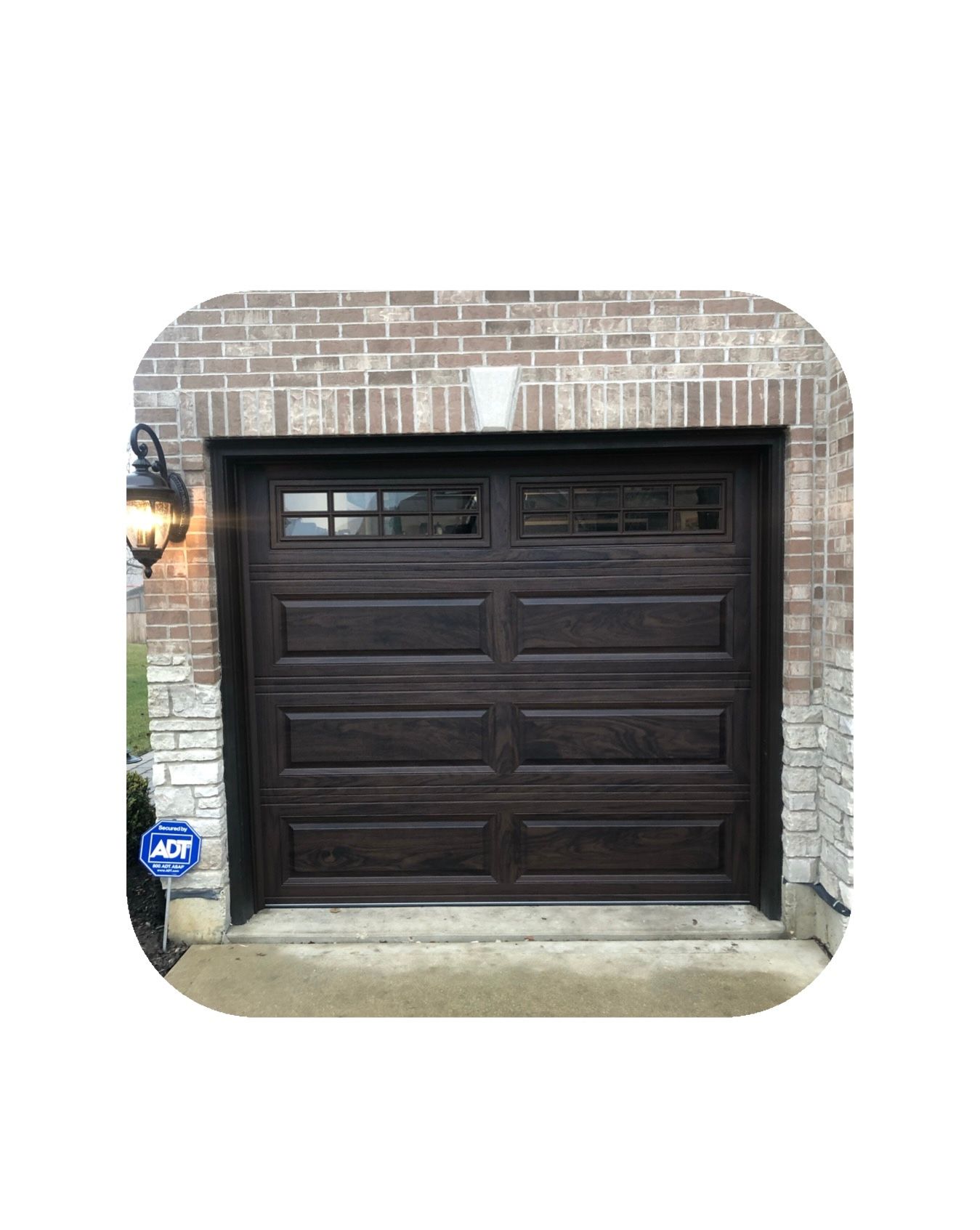 Dark brown garage door with window panels, framed by brick and stone, with a side lamp.