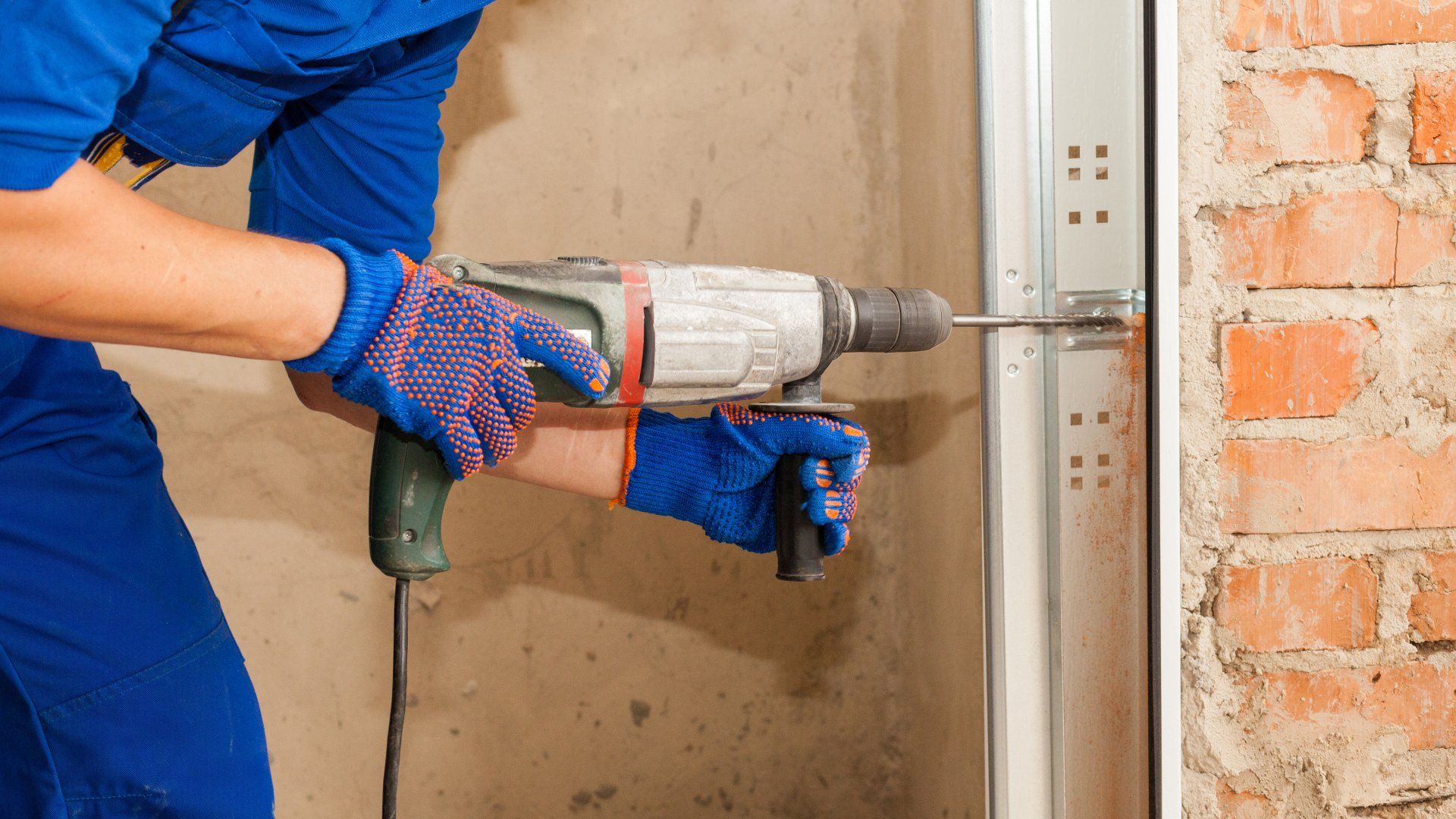 Panoramic and cropped view of professional worker holding electric drills in hands, making hole in brick copy space wall, install metal rail on garage door system