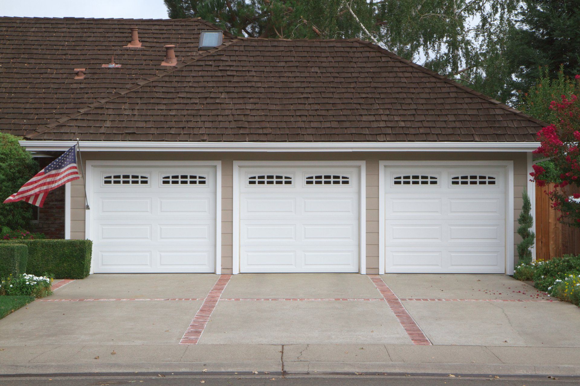 Three white garage doors with windows, brown roof, American flag, brick driveway accents.