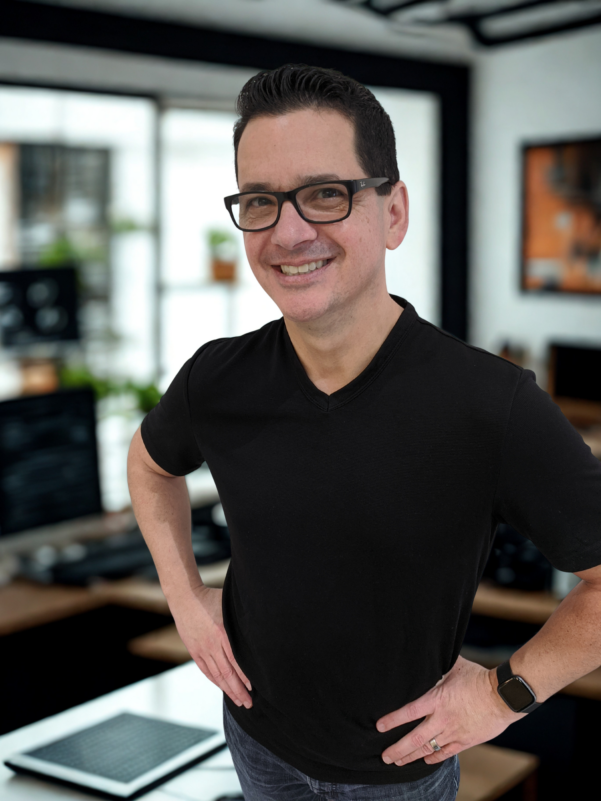 Man with glasses and a black v-neck smiles with hands on hips in an office setting.