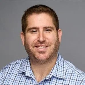 A smiling man with brown hair and a light beard wearing a blue plaid shirt in a studio setting.