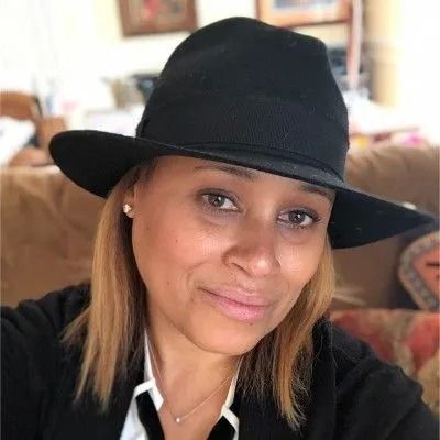 Woman with light brown skin wearing a black fedora, smiling, in a home setting.