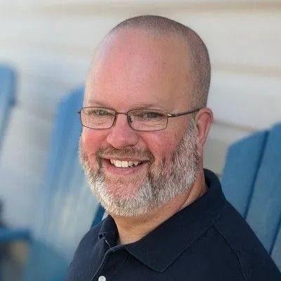 Man with glasses and a beard smiles, seated outdoors in front of blue chairs.