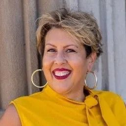 Woman with blonde hair, wearing a yellow top and large gold hoop earrings, smiles at the camera.
