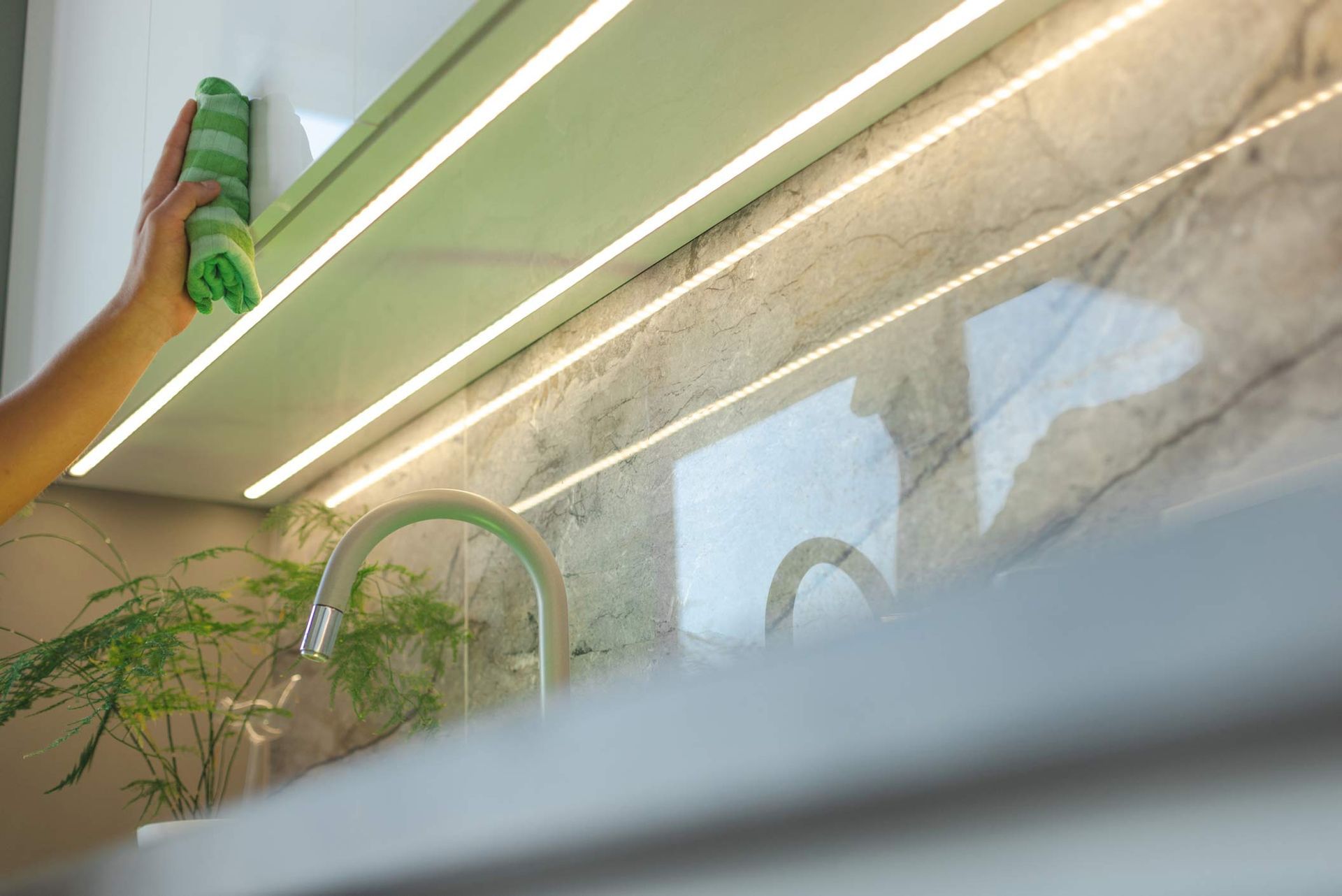 A person cleaning a kitchen cabinet, illuminated by LED, showcasing a polished marble wall.
