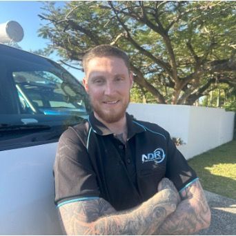 Man with tattoos in black polo shirt smiles next to white van. — NDR Trade Solutions in Robina, QLD