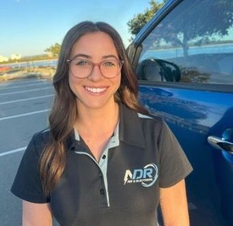 Woman with glasses, smiling, wearing a black polo shirt in front of a blue car. — NDR Trade Solutions in Robina, QLD