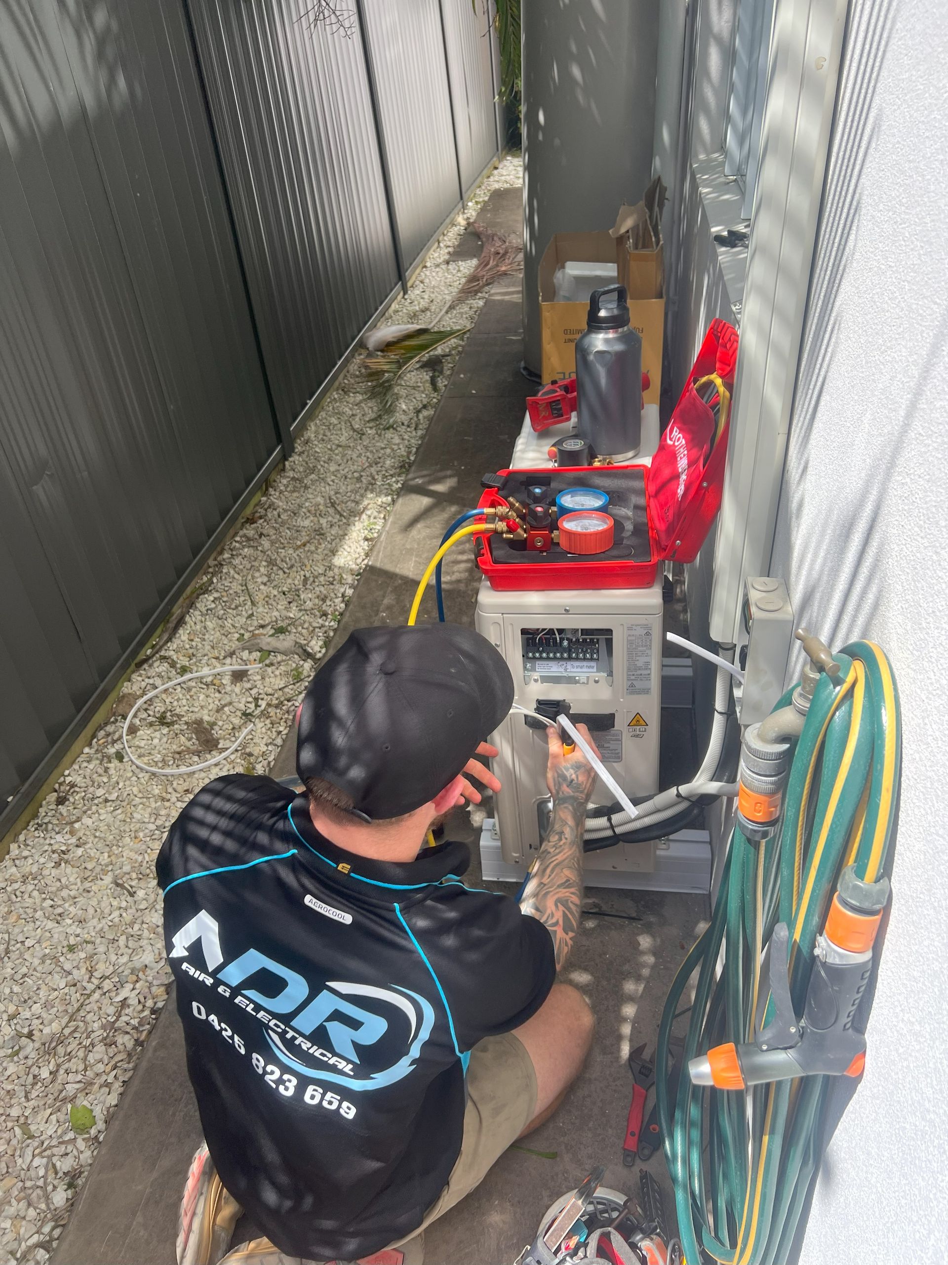 A Man in Black Shirt Works on Electrical Equipment Outdoors Next to a Wall — NDR Trade Solutions in Robina, QLD