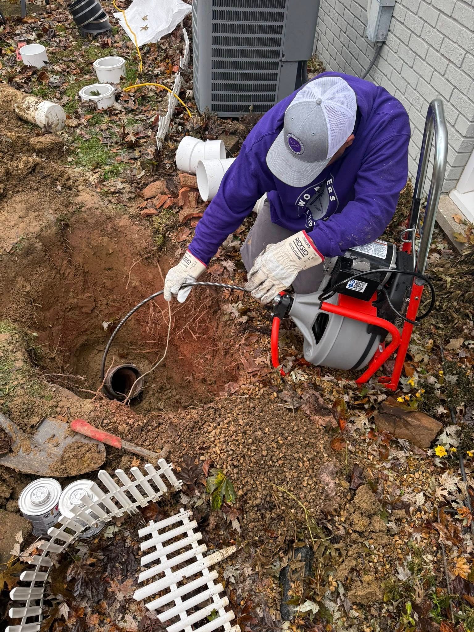 A person in a purple shirt and gray cap uses a sewer drain cleaning machine to work on a pipe inside an outdoor pit.