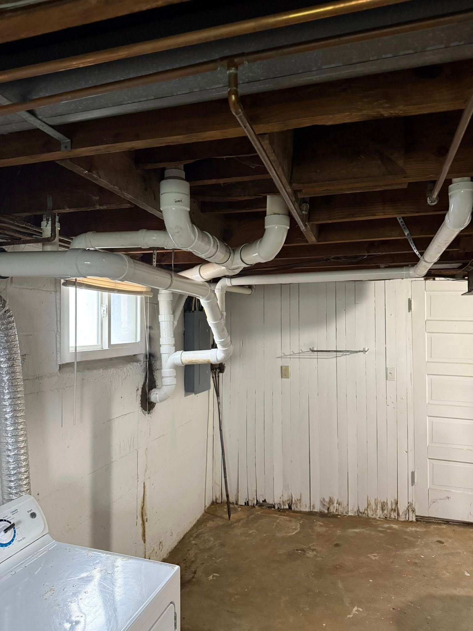 White PVC plumbing pipes running along the wooden floor joists and concrete wall in a basement.