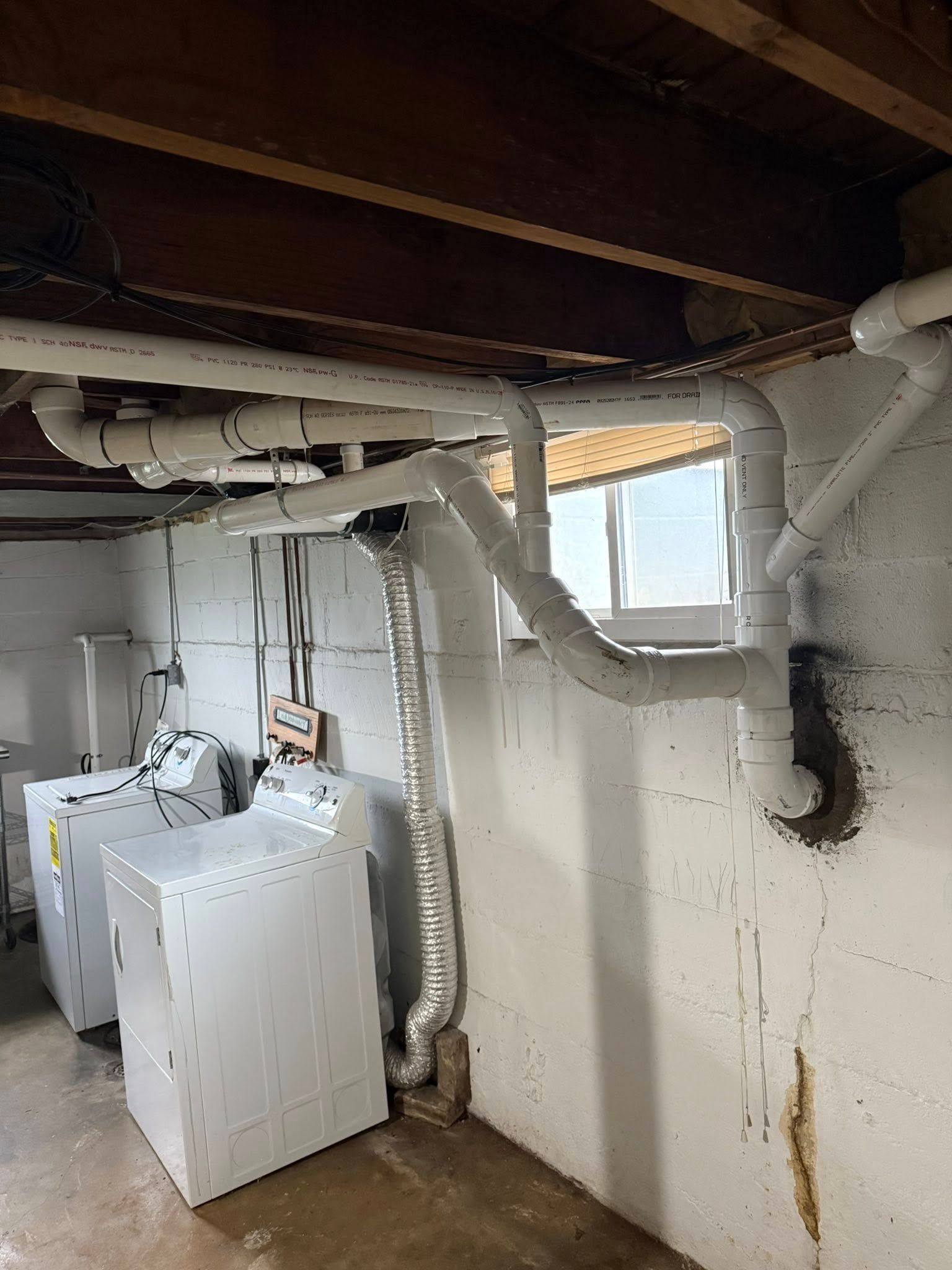 A laundry room basement featuring a clothes dryer connected to a silver vent pipe and white PVC plumbing along the ceiling.