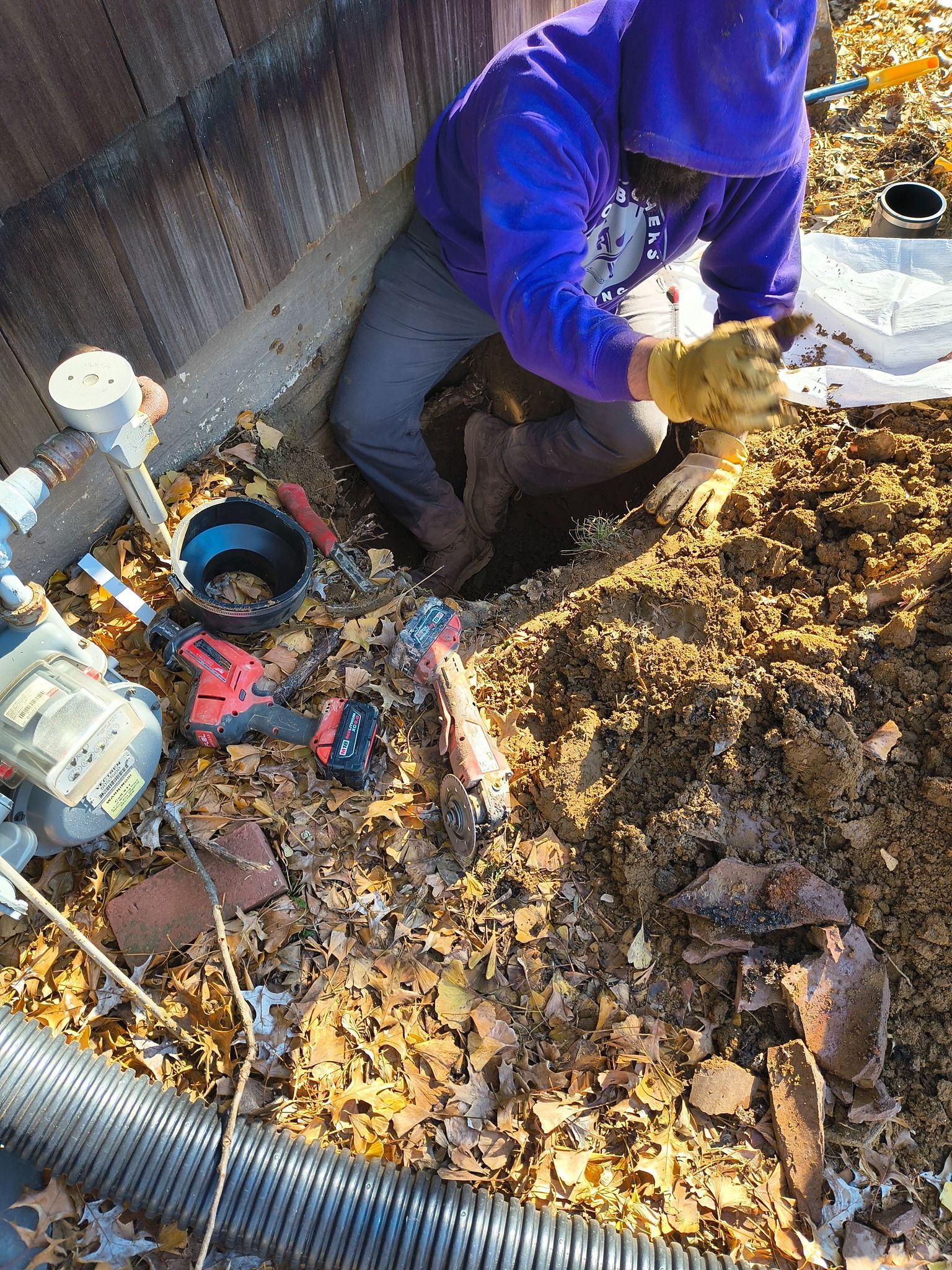 A person in a purple hoodie and work gloves kneels in a leaf-covered yard, working on underground utility equipment.