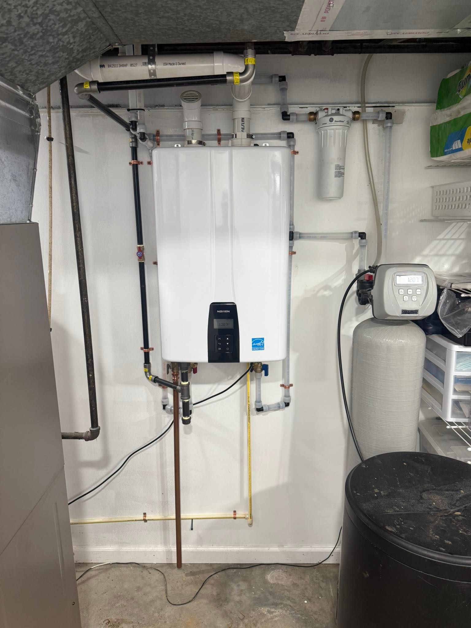 A white tankless water heater mounted on a wall next to a water softening system in a basement setting.