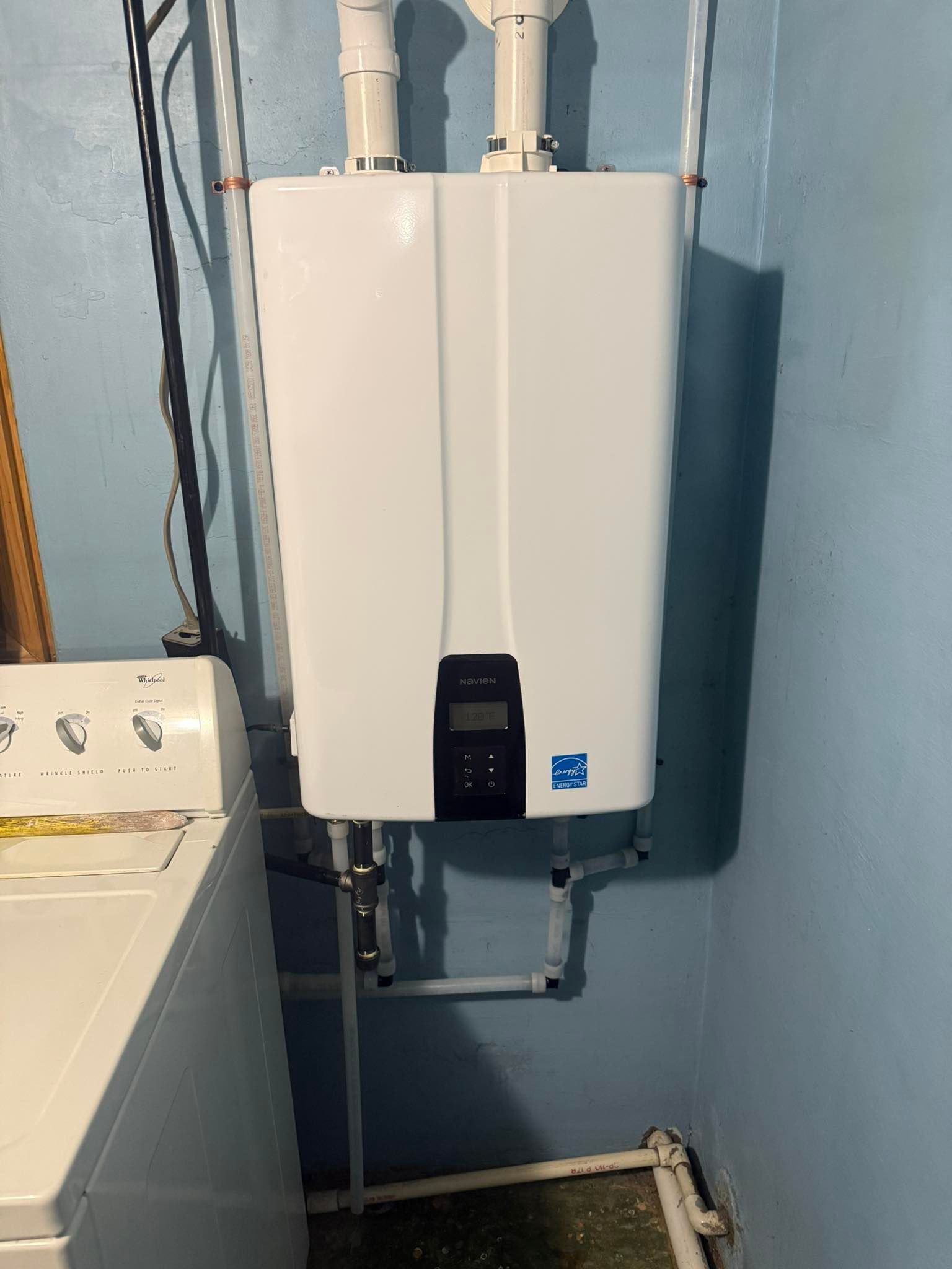 A white tankless water heater mounted on a blue laundry room wall next to a washing machine.