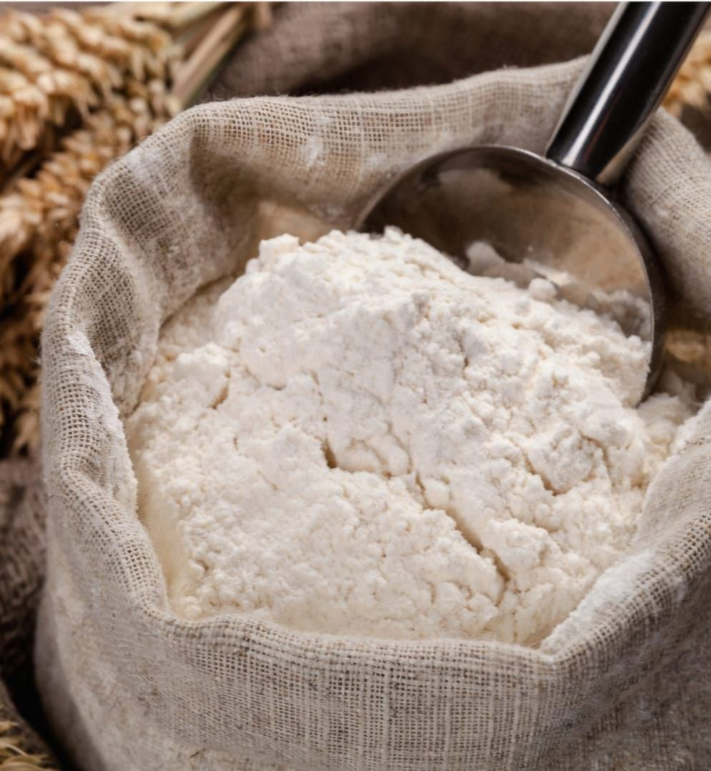 Una cuchara de metal reposa dentro de un saco de arpillera lleno de harina blanca, con tallos de trigo al fondo.