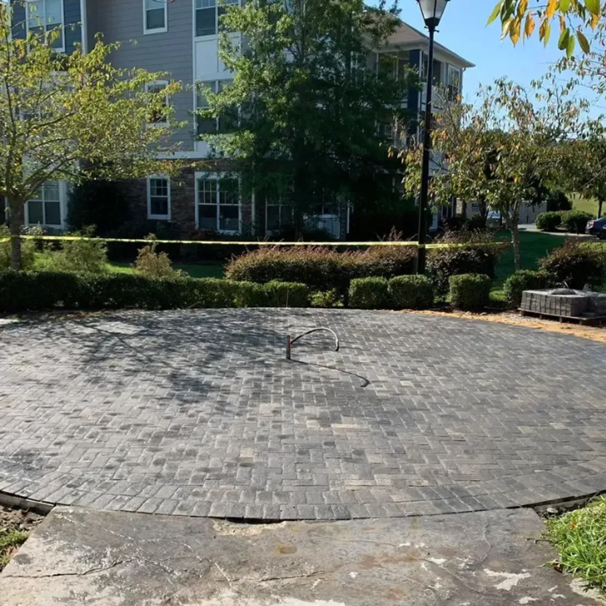 Circular brick patio in front of apartment building, bordered by bushes and trees; a light post is in the background.