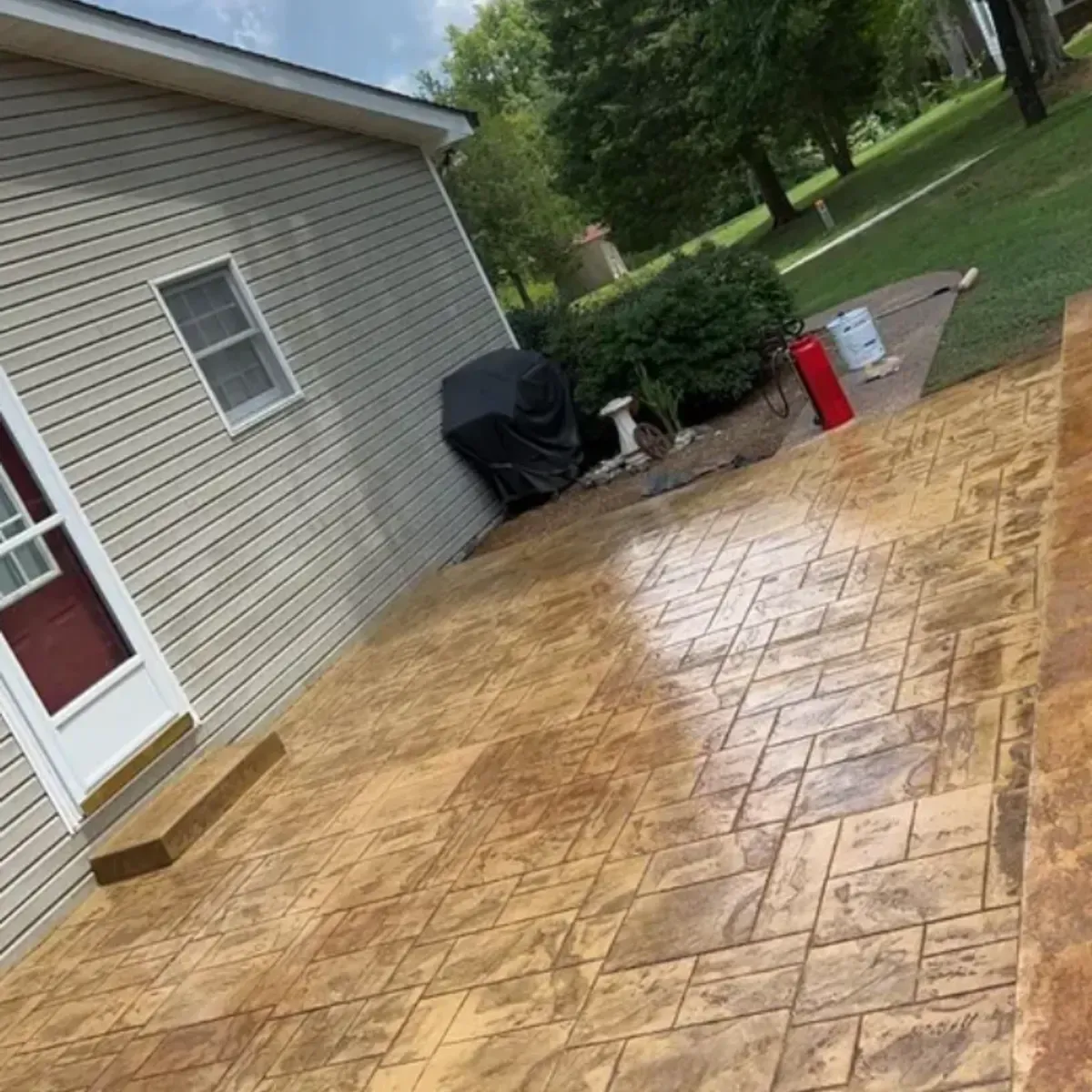 Tan stamped concrete patio next to a house with a grill, trees, and a red fire extinguisher.