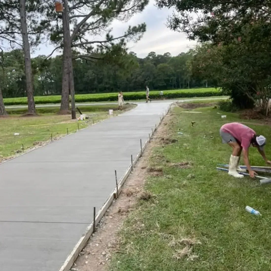 Man working on new concrete driveway outdoors; green grass, trees in background.