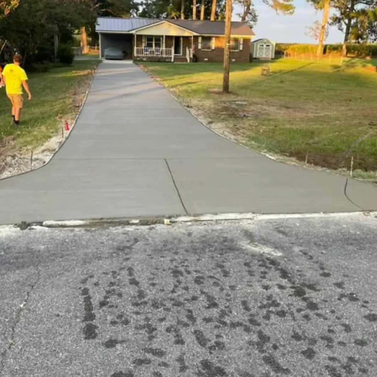 Newly poured concrete driveway leading to a house with a person in a yellow shirt walking on the grass.