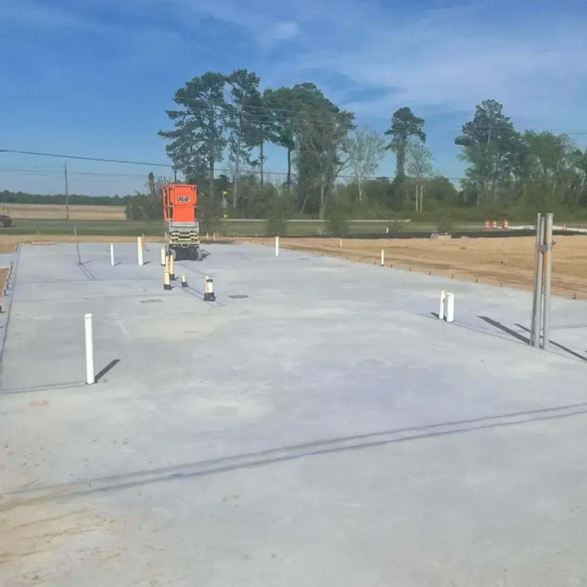 Newly poured concrete foundation with plumbing pipes, construction site with trees and lift.