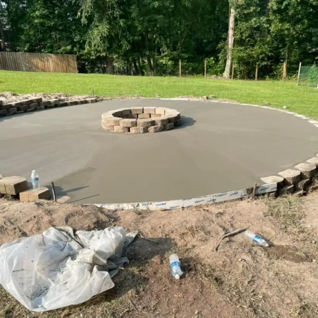 Circular concrete patio with a stone fire pit, surrounded by landscaping blocks, in a backyard.