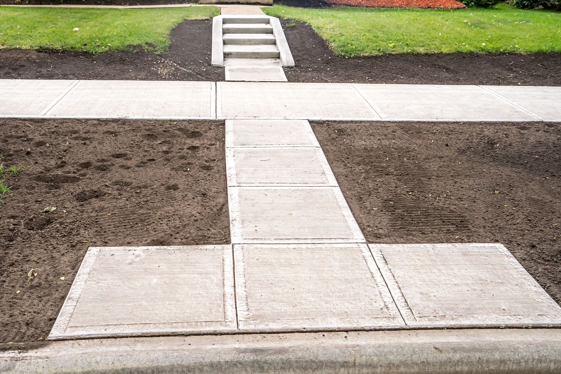 Concrete walkway leading to stairs up to a house, surrounded by dirt and grass.