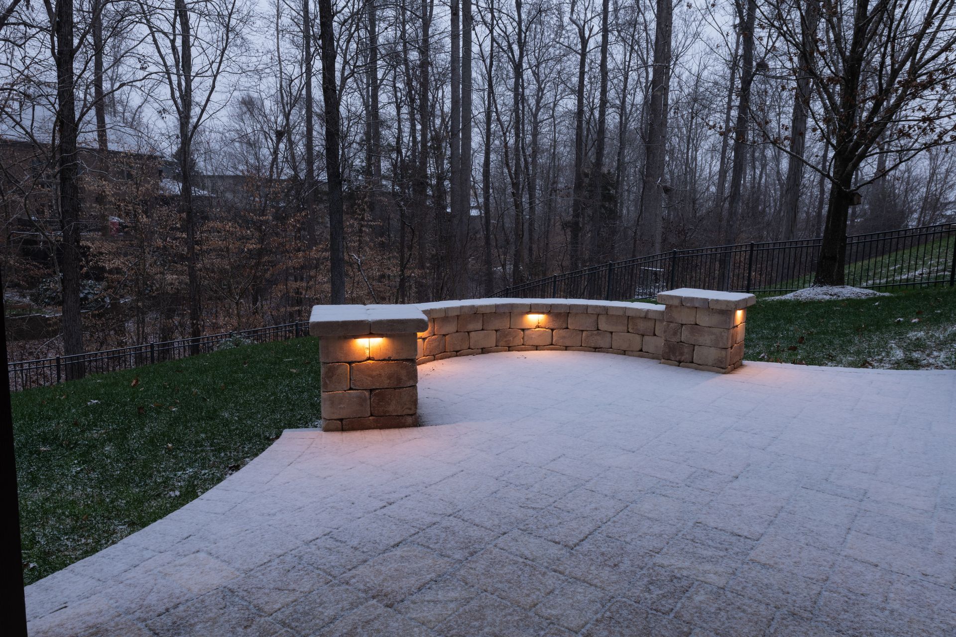 Snowy patio with illuminated stone wall and trees in the background.