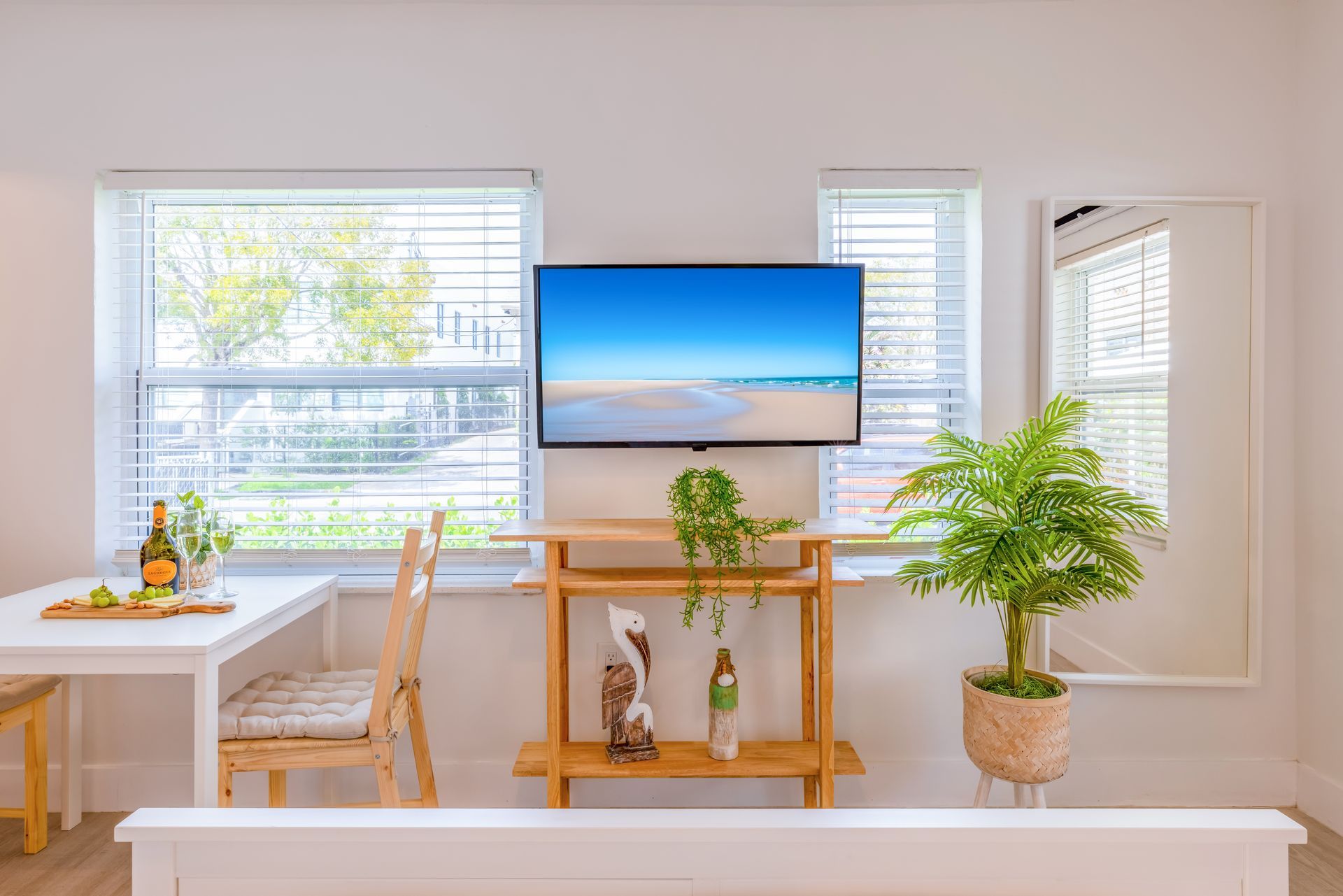 Bright living room with a TV mounted on a wooden shelf, near windows. Fern and plants. White walls and furniture.