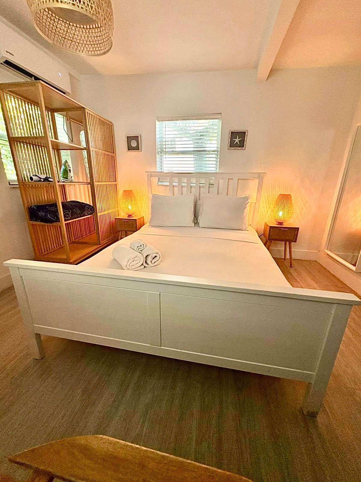 White bedroom with bed, nightstands, shelving unit, window, and mirror.