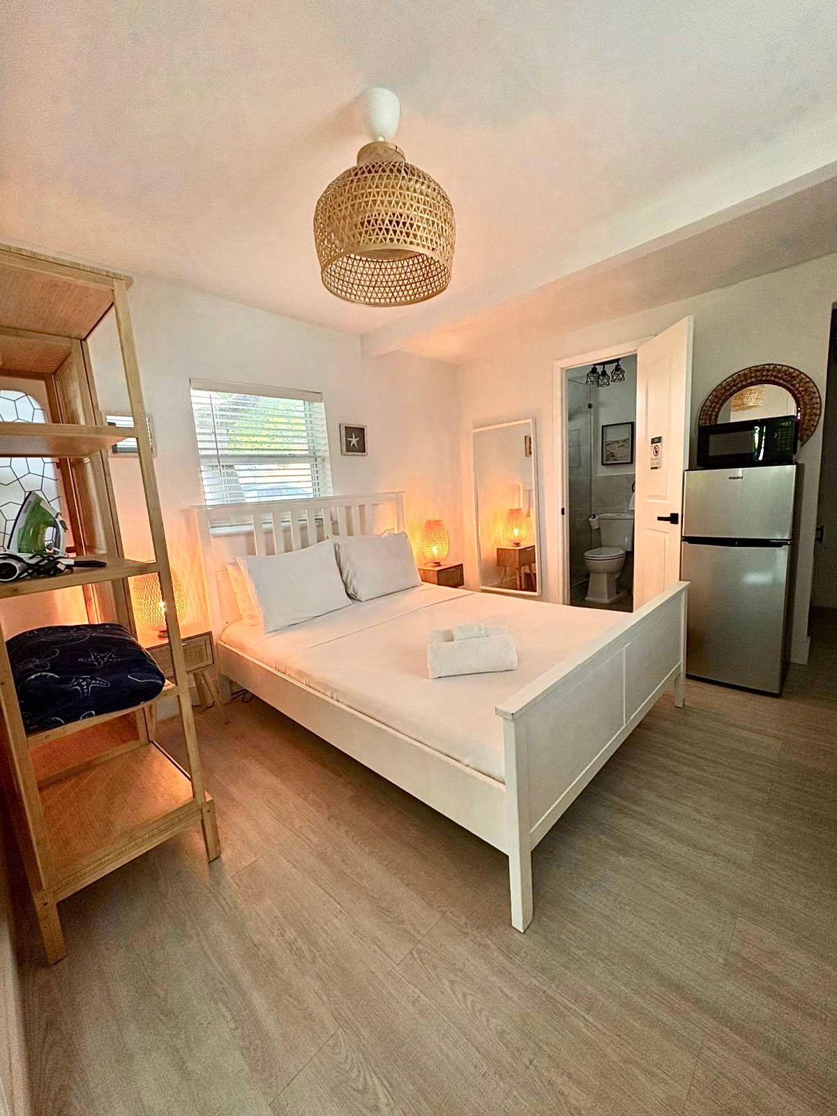Bedroom with a white bed, wood shelves, and a refrigerator. Warm lighting and a woven lampshade.