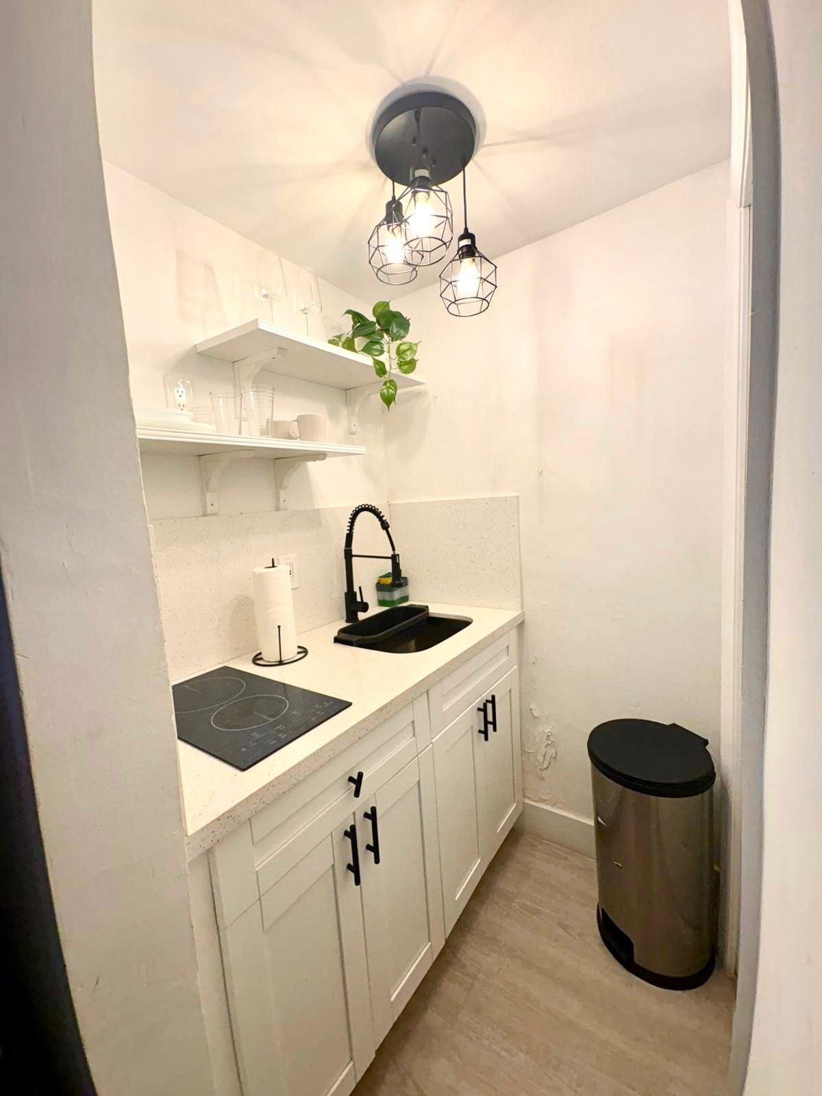 Small white kitchen with black sink, stove, and cabinet handles. Overhead lights, shelves, and trash can.