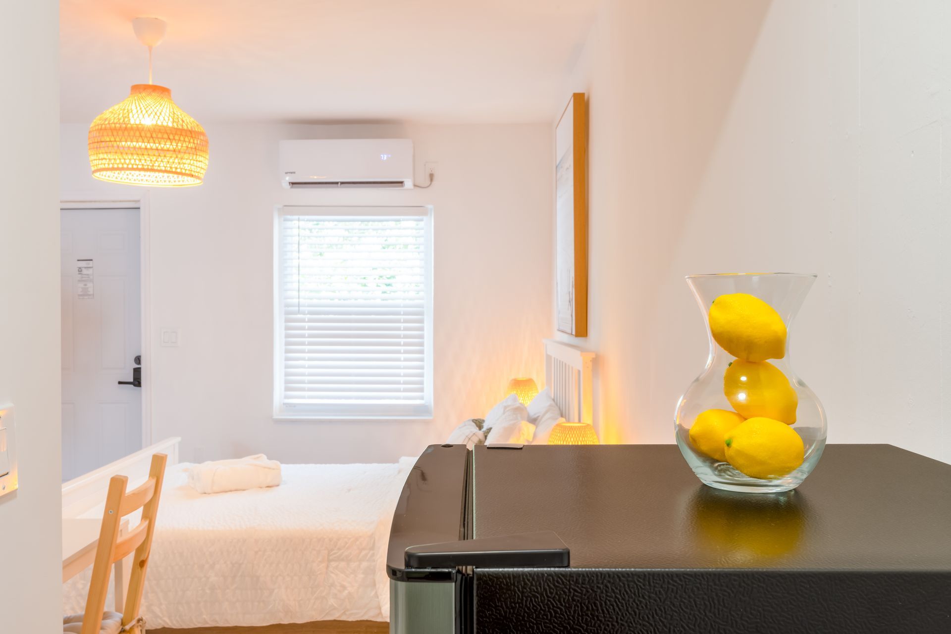 Small, white room with bed, fridge, lemons in a vase, and a window with blinds.