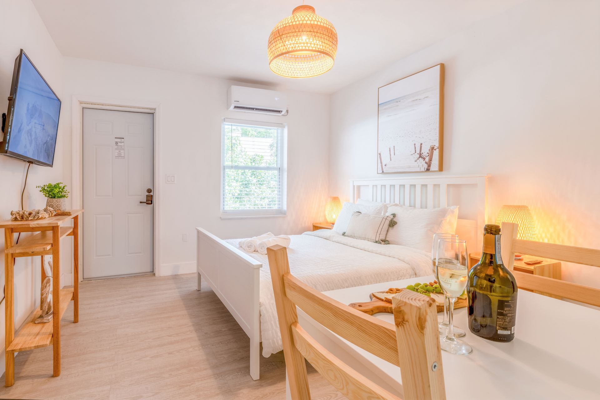 White bedroom with bed, table set for a meal, TV, and window, lit by a woven light fixture.