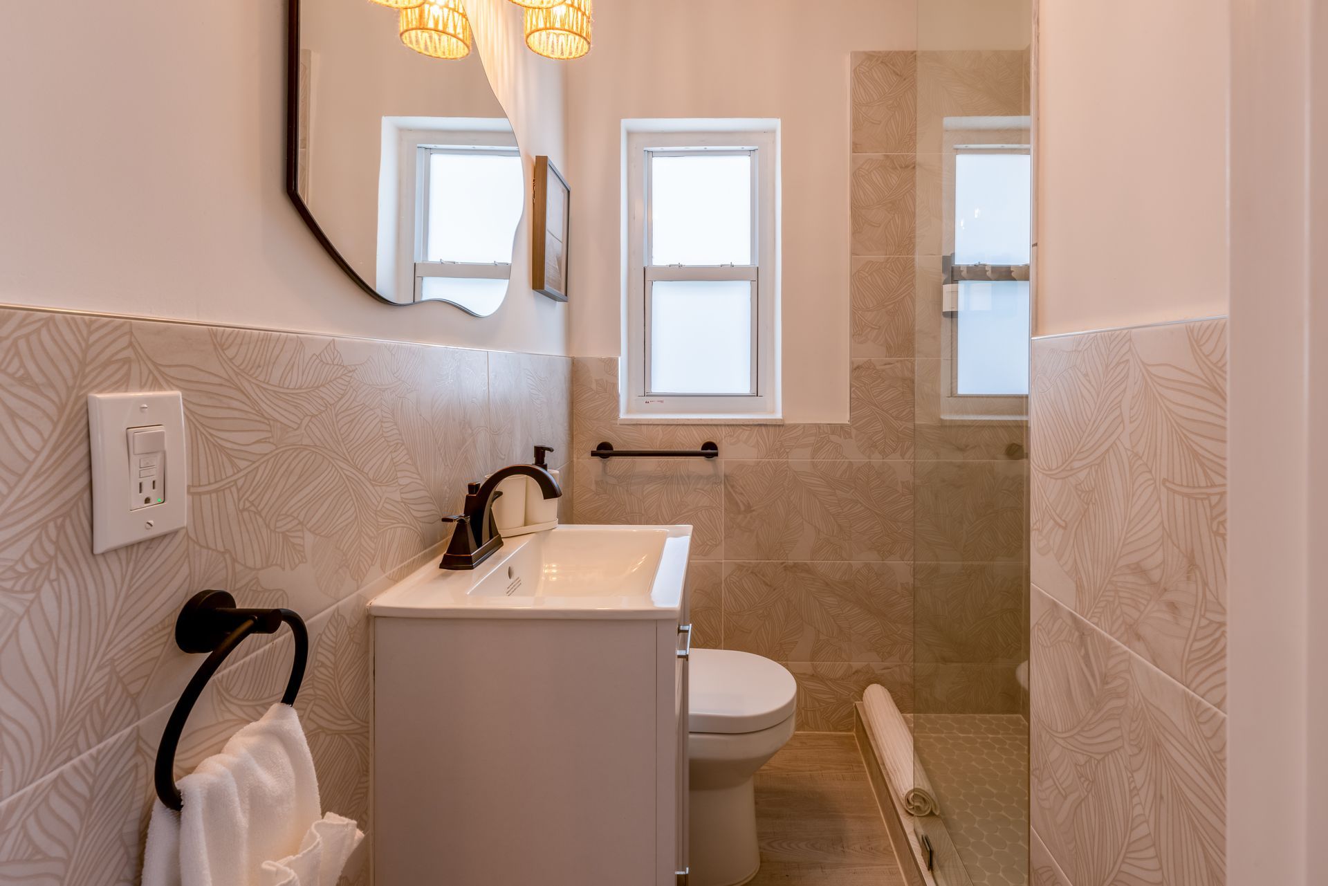 Small beige and white bathroom with sink, toilet, and shower behind glass.