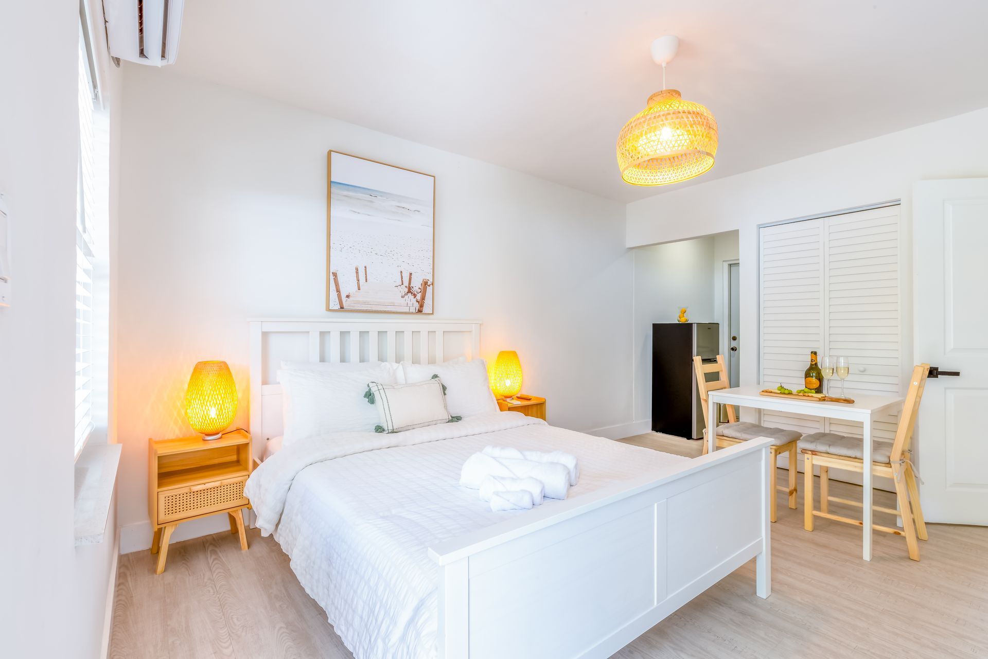 Bright white bedroom with a bed, two nightstands, a dining table, and a framed beach scene.
